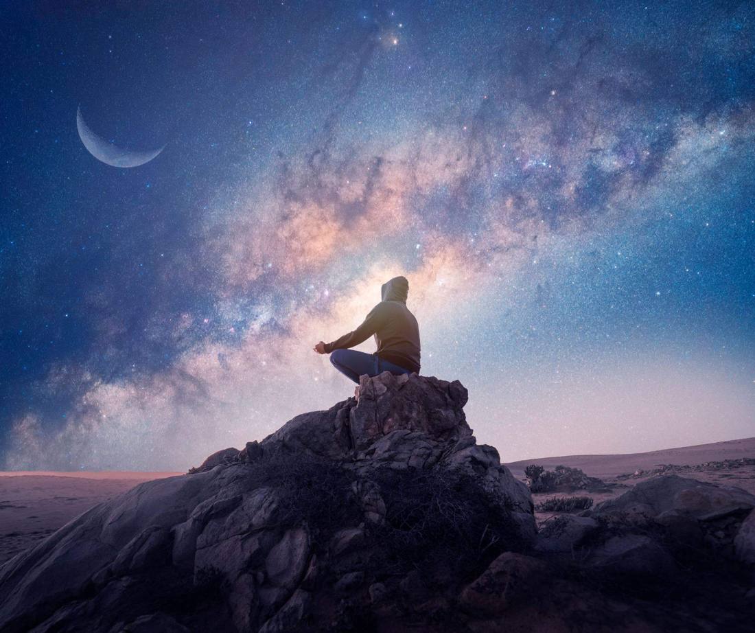 Persona meditando sobre una roca bajo la Vía Láctea y la luna creciente
