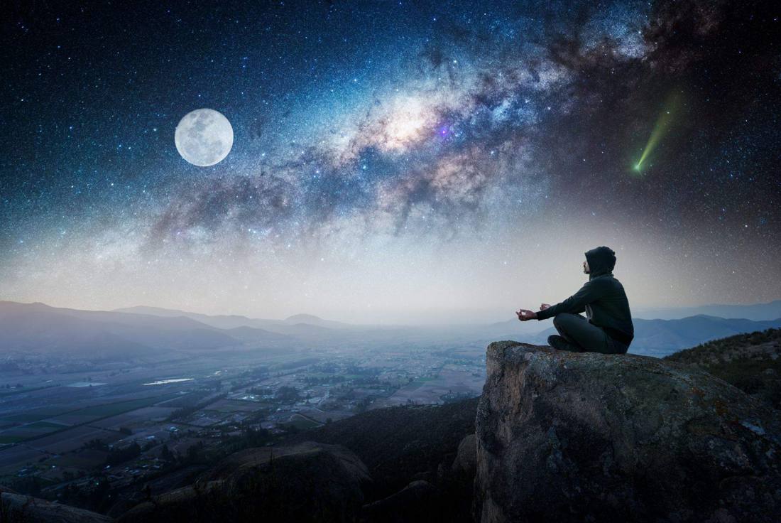 Una persona sentada en una roca, en calma, mirando la Vía Láctea y la luna sobre el valle